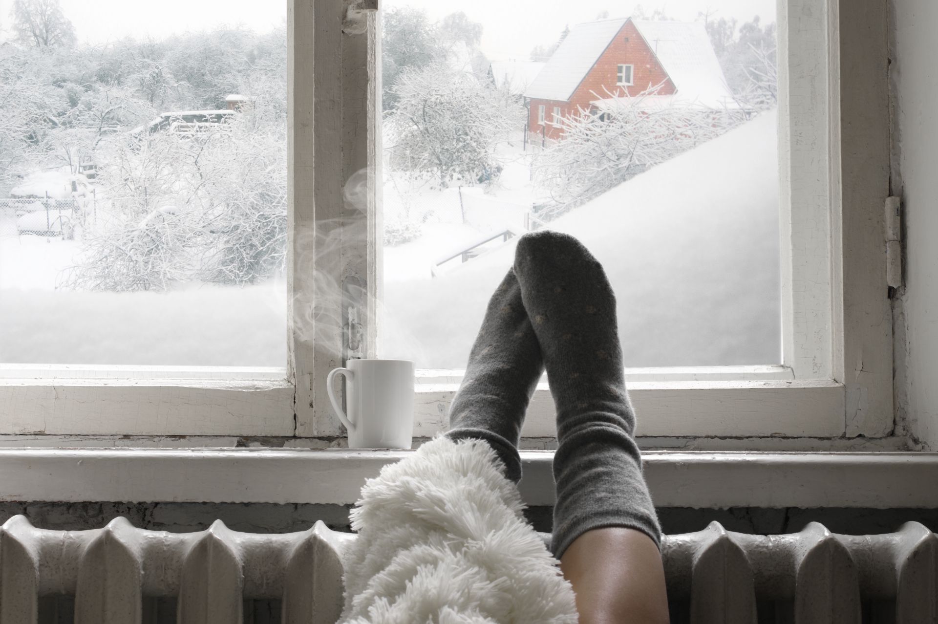 Person in wool socks relaxing at snowy window with mug and red barn visible outside